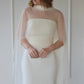 Woman wearing a white wedding dress with a sheer overlay against a plain background